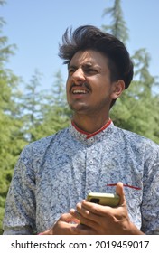 Closeup Portrait Of A Indian Good Looking Young Guy Standing Outdoor In Nature And Holding Mobile Phone In His Hands With Looking Sideways 