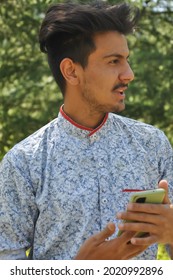 Closeup Portrait Of A Indian Good Looking Young Guy Standing Outdoor In Nature And Holding Mobile Phone In His Hands With Looking Sideways 