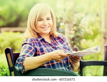 Closeup Portrait Of Joyful Blond Mature Woman Reading Magazine In Garden On Summer Day
