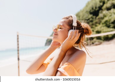 Close-up portrait of joyful tanned woman relaxing with favorite music at beach. Outdoor shot of smiling female model in headphones spending time at resort. - Powered by Shutterstock