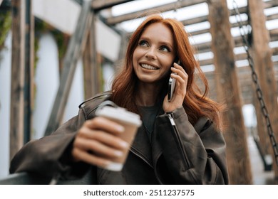 Closeup portrait of joyful young woman with long red hair enjoying coffee on city swing, happily chatting on mobile phone outdoors in city park. Happy red-haired female enjoy coffee on park swing. - Powered by Shutterstock