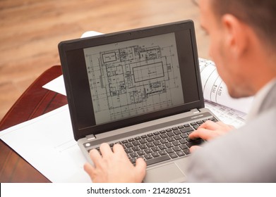 Close-up portrait of laptop with blueprints, architect sitting from behind working on architectural plan, interior shot