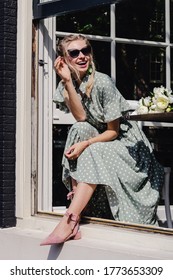 Close-up portrait of laughing blonde girl in pale green long dress with sunglasses, nice hair and beautiful shoes with ankle ties looking out of window of the city cafe. Street style look
