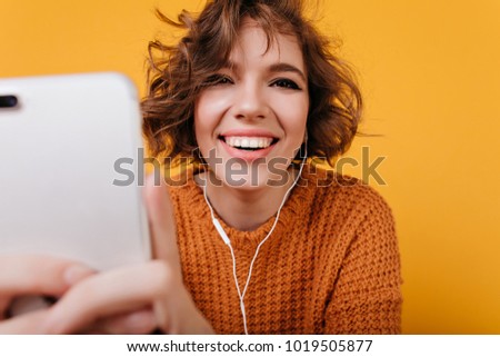 Close-up portrait of laughing white girl using phone for selfie. Studio shot of young woman with wavy hair listening music and posing with new smartphone.