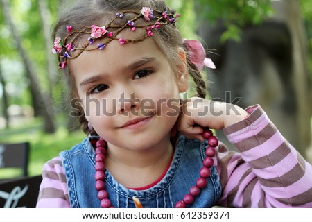Close-up portrait of little beautiful blonde girl wearing an elegant wreath of leaves and flowers posing in a green meadow. Fashionable attractive child, gorgeous fair hair female, outdoor summer shot