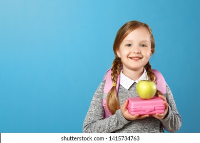 Closeup Portrait Of A Little Girl Schoolgirl On A Blue Background. The Child Has A Satchel Behind His Back And Holds A Lunchbox In His Hands. Back To School. The Concept Of Education.