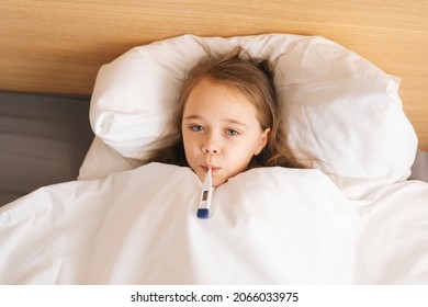 Close-up portrait of little sick girl lying in bed measuring temperature with thermometer in mouth at home, looking at camera. Unhappy child with high fever lying at bad in medical clinic.