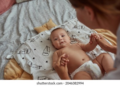 Closeup Portrait Of Lovely Baby In Diaper Without Clothes, Lying On Back, Looking With Curiosity At Camera. Unrecognised Woman Holding Baby's Hands.