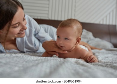 Closeup portrait lovely mom and sweet baby on bed. Playful smiling mother tickling baby's chin. Baby routine tummy time on bed with mother engaging to stay on belly longer.