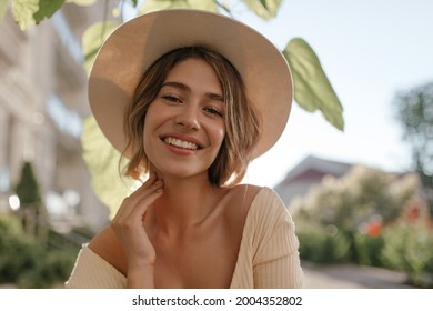 Close-up portrait of lovely white female model with natural make-up expressing good mood. Lovely fair woman in stylish hat holds her chin with her hand.