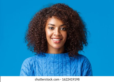 Close-up portrait lovely young african-american woman with curly, afro haircut, smiling and looking camera with happy pleasant expression, enjoying winter holidays, wearing sweater, blue background - Powered by Shutterstock