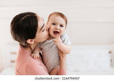 Closeup portrait of loving young mom kissing little kid in cheek, bonding with her adorable son or daughter, sitting on bed at home, free space - Powered by Shutterstock