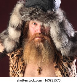 Close-up Portrait Of A Man With A Long Beard And Mustache, Wearing A Fur Hat, Studio Shot On A Burgundy Background