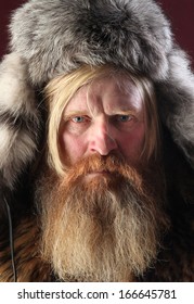 Close-up Portrait Of A Man With A Long Beard And Mustache, Wearing A Fur Hat, Studio Shot On A Burgundy Background