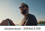 Close-up portrait of a man standing on a lake pier at sunset, leaning on a waterboard. Male surfer holding a short surfboard while standing on a pier with a motorboat in the background