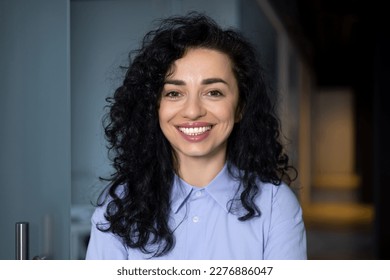 Close-up portrait of mature adult business woman, boss smiling and looking at camera, pleased hispanic woman with curly hair in blue shirt inside office at workplace in hall corridor.