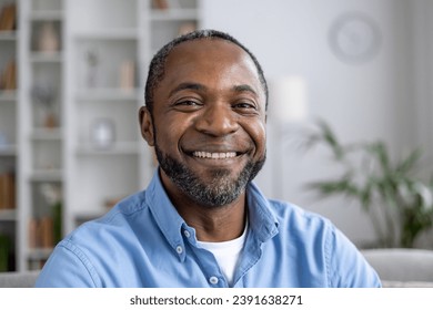 Close-up portrait of mature experienced African American man smiling and looking at camera while sitting on sofa in living room at home. - Powered by Shutterstock