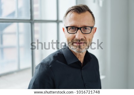 Close-up portrait of a middle-aged man wearing black shirt and eyeglasses while looking at camera with confidence in the office