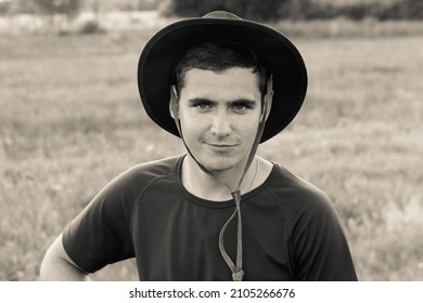 Closeup Portrait Of Millennial Man With Cowboy Hat, Standing On Nature Background, Outdoors. Countryman, Rancher On Farmland. Sepia, Black And White. Retro, Vintage.