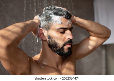 Closeup Portrait Of Muscular Bearded Man Washing Hair And Body Using Male Shampoo Taking Shower With Foam Standing Under Hot Water In Modern Bathroom At Home. Men's Beauty Routine And Hygiene Concept