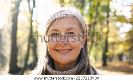 Close-up portrait of naturally beautiful senior cute gray-haired woman standing in park in autumn wearing coat looking at camera smiling. Pretty mature kind good-looking female on fresh air.