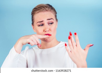 Close-up Portrait Of Nervous Unhappy Young Blonde Woman Looking At A Broken Fingernail And Crying . Red Long Nails Manicure Broken Nail Blue Background In Studio