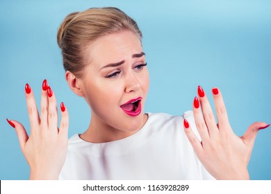 Close-up Portrait Of Nervous Unhappy Young Blonde Woman Looking At A Broken Fingernail And Crying . Red Long Nails Manicure Broken Nail Blue Background In Studio