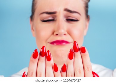 Close-up Portrait Of Nervous Unhappy Young Blonde Woman Looking At A Broken Fingernail And Crying . Red Long Nails Manicure Broken Nail Blue Background In Studio