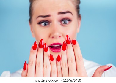 Close-up Portrait Of Nervous Unhappy Young Blonde Woman Looking At A Broken Fingernail And Crying . Red Long Nails Manicure Broken Nail Blue Background In Studio