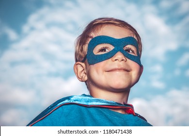 Closeup Portrait Of A Nice Little Superhero Over Blue Sky Background, A Cheerful Child Wearing Superman Mask And Cape, Happy Childhood Concept