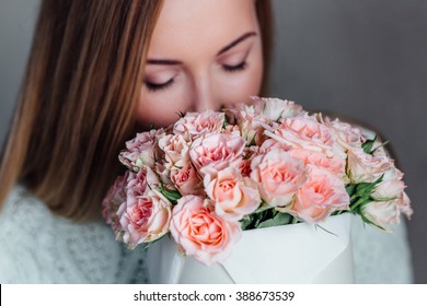 Closeup portrait Nice young blonde girl with Blue Eyes and healthy Long Blond Hair holding roses bouquet in hat box  against the  plastered wall, wearing jeans and knit sweater, smelling flowers.