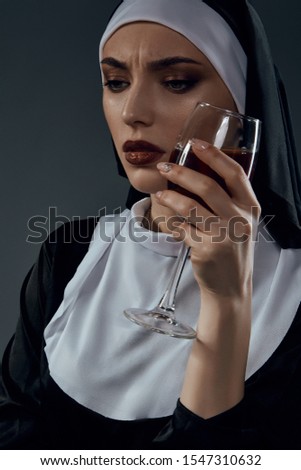 Close-up portrait of a nun, posing on a black background. She's wearing dark nun's clothing. The nun is holding a glass of wine in her left hand at her face and looking down. 
