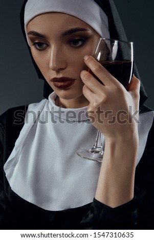 Close-up portrait of a nun, posing on a black background. She's wearing dark nun's clothing. The nun is holding a glass of wine in her left hand at her face and looking down.  
