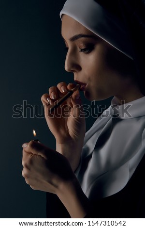 Close-up portrait of a nun, taking on a black background. She wearing dark nun's clothing. The nun is lighting a cigarette. She looking down. 