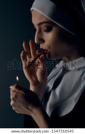Close-up portrait of a nun, taking on a black background. She wearing dark nun's clothing. The nun is lighting a cigarette. She looking down.