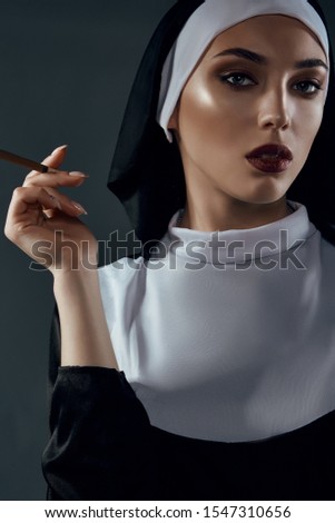 Close-up portrait of a nun, taking on a black background. She wearing dark nun's clothing. The nun is holding a cigarette in right hand and looking straight.