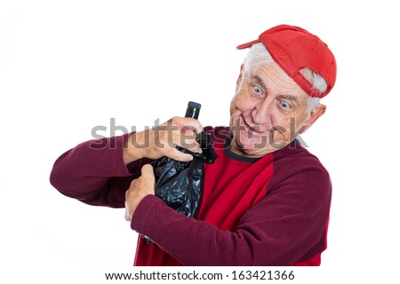 Closeup portrait of an old guy, senior grandfather, elderly man wearing red shirt and cap, really drunk holding bottle of alcohol in black bag in hands, isolated on white background. Facial expression