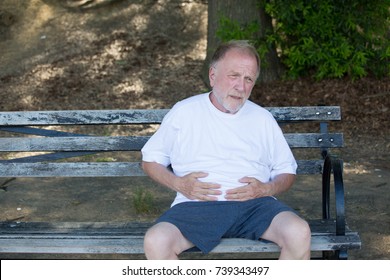 Closeup Portrait, Old Man Clutching Stomach, Having Belly Pain, Sitting On Bench, Isolated Outdoors, Green Trees Background. Full After A Meal Or Pain After Walking