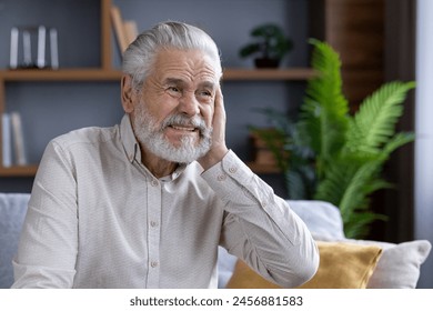 Close-up portrait of an older man suffering from severe ear pain, sitting on the sofa at home and grimacing, holding his hand behind his head. - Powered by Shutterstock