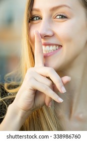 Closeup portrait of one beautiful happy smiling blonde young woman with long hair outdoor looking forward with hush gesture on blurred background, vertical picture - Shutterstock ID 338986760