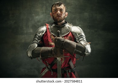 Close-up. Portrait of one brutal bearded man, medeival warrior or knight with dirty wounded face holding big sword isolated over dark background.