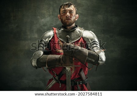 Close-up. Portrait of one brutal bearded man, medeival warrior or knight with dirty wounded face holding big sword isolated over dark background.