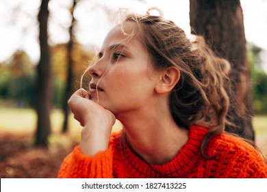 Closeup portrait of a pensive woman looking away, wearing an orange knitted sweater posing on a fall nature background. The beautiful female has a thoughtful expression, resting outdoor in the park.