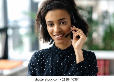 Close-up portrait of pleasant charming young mixed race curly haired woman in headset and stylish shirt, call center worker or support operator, looking directly at camera, smiling friendly - Shutterstock ID 2118440384