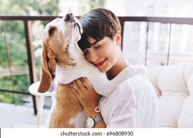 Close-up portrait of pleased girl with short brown hair embracing funny beagle dog with eyes closed. Smiling young woman in white shirt enjoying good day and posing with pet on terrace.