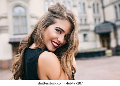 Closeup portrait of pretty girl with long curly hair smiling to camera in city on old building background. She wears black dress, red lips. View from back. - Powered by Shutterstock