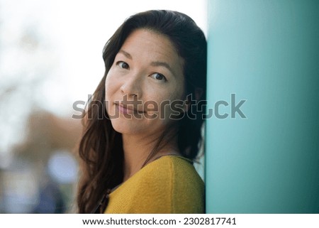 Close-up portrait of pretty young asian woman leaning to a wall outside and staring at camera
