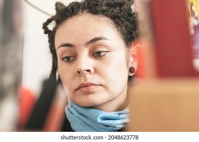 Closeup Portrait Of Pretty Young White Girl Hispanic Hispanic Latin Art Director, With Beautiful Eyes, Blue Bandanna And Dreadlocks Looking At Her Working Tools On Her Table, To Choose One.