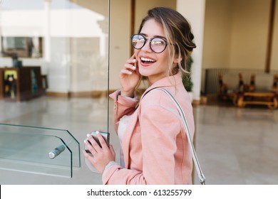 Closeup Portrait Of Pretty Young Woman With Hair Up Entering Building, Glass Door Into Office, Hotel, Business Centre. Wearing Fashionable Glasses, Gray Pants, Pink Jacket.