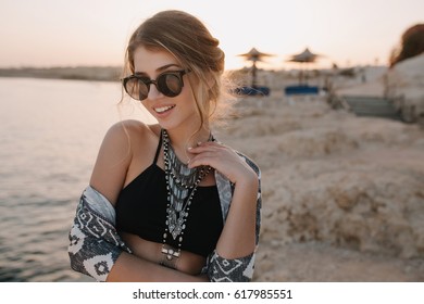 Closeup Portrait Of Pretty Young Woman On Sunset, On Beach With Sensual Look. Wearing Trendy Black Top, Stylish Sunglasses, Necklace, Cardigan, Cape With Ornaments.